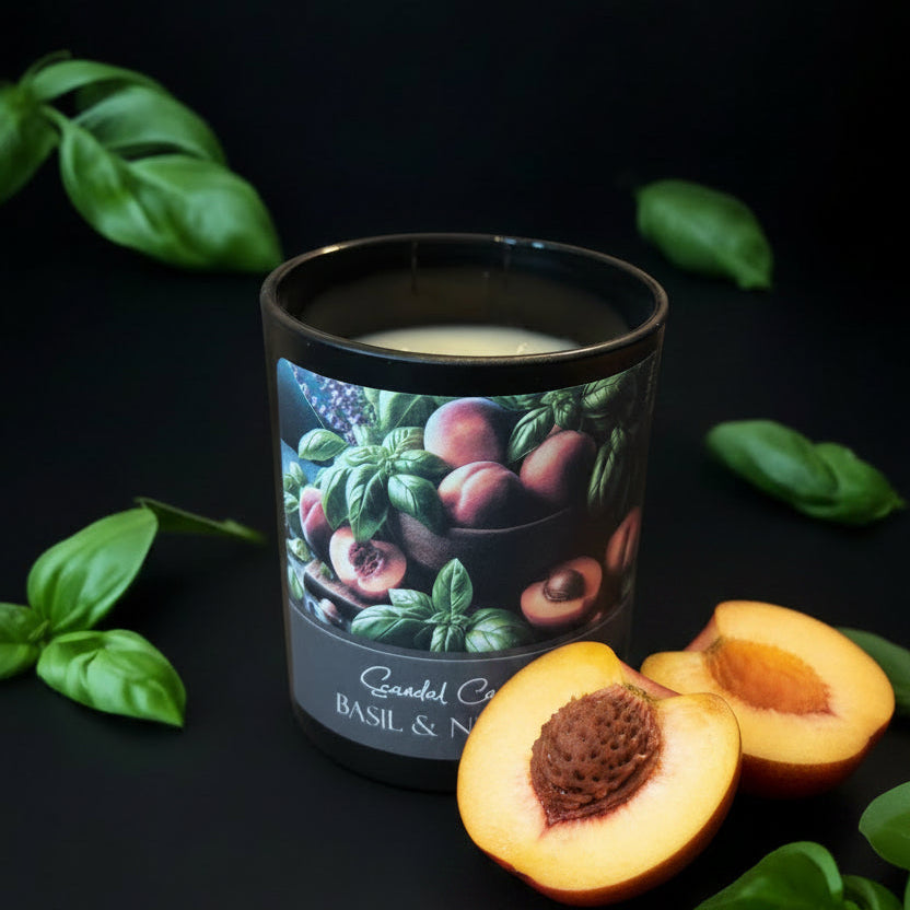 A candle in a dark glass jar with a printed label showing basil leaves and nectarines, styled on a dark surface with scattered green basil leaves. The label reads 'Scandal Candle Co' and 'BASIL & NECTARIN'.