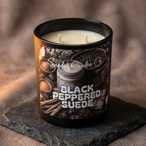 A single-wick candle in a black glass jar with cream-colored wax, resting on a dark slate coaster against a textured brown fabric background. The label features illustrations of spices and the text 'Scandal Candles Co' and 'BLACK PEPPERED SUEDE'.
