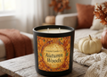 A black scented candle labeled 'Autumn Woods' by Scandal Candles Co. sits on a rustic wooden table in a cozy living room setting with autumn-themed decor, including pumpkins, a knitted blanket, and a couch in the background.