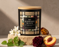 A candle tin labeled 'MY BURBERRY BLACK' by Scents of Candle Co., styled on a stone pedestal with white flowers, mint leaves, a rose, a peach half, and amber stones against a warm beige background.